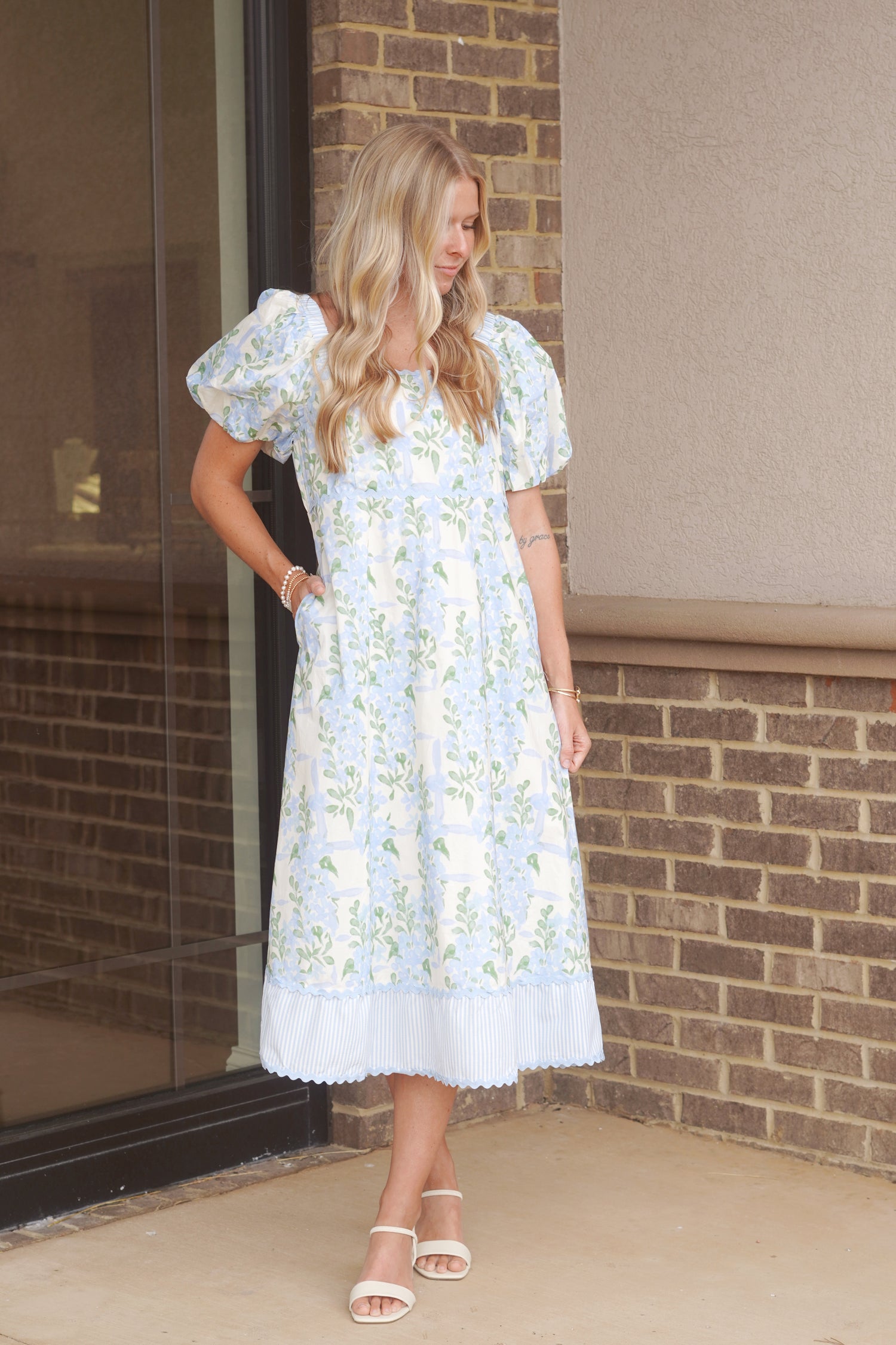 Women wearing blue floral print dress with a blue and white stripe hem detail in the bottom of the dress.  This dress also has puff sleeves and a square neckline. 