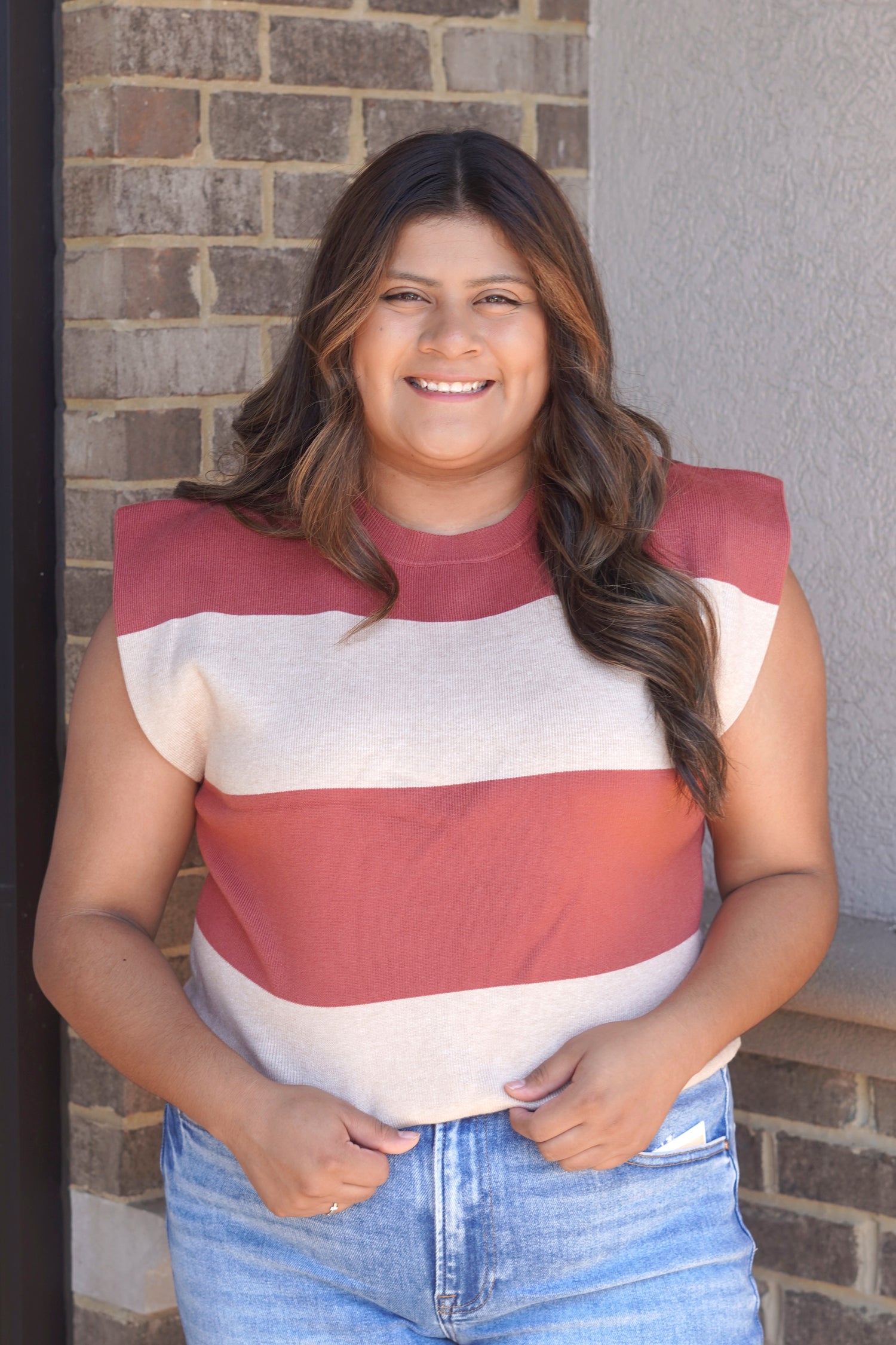 A  woman wearing a sleeveless knit top with wide horizontal stripes in dusty rose and cream, paired with blue jeans.