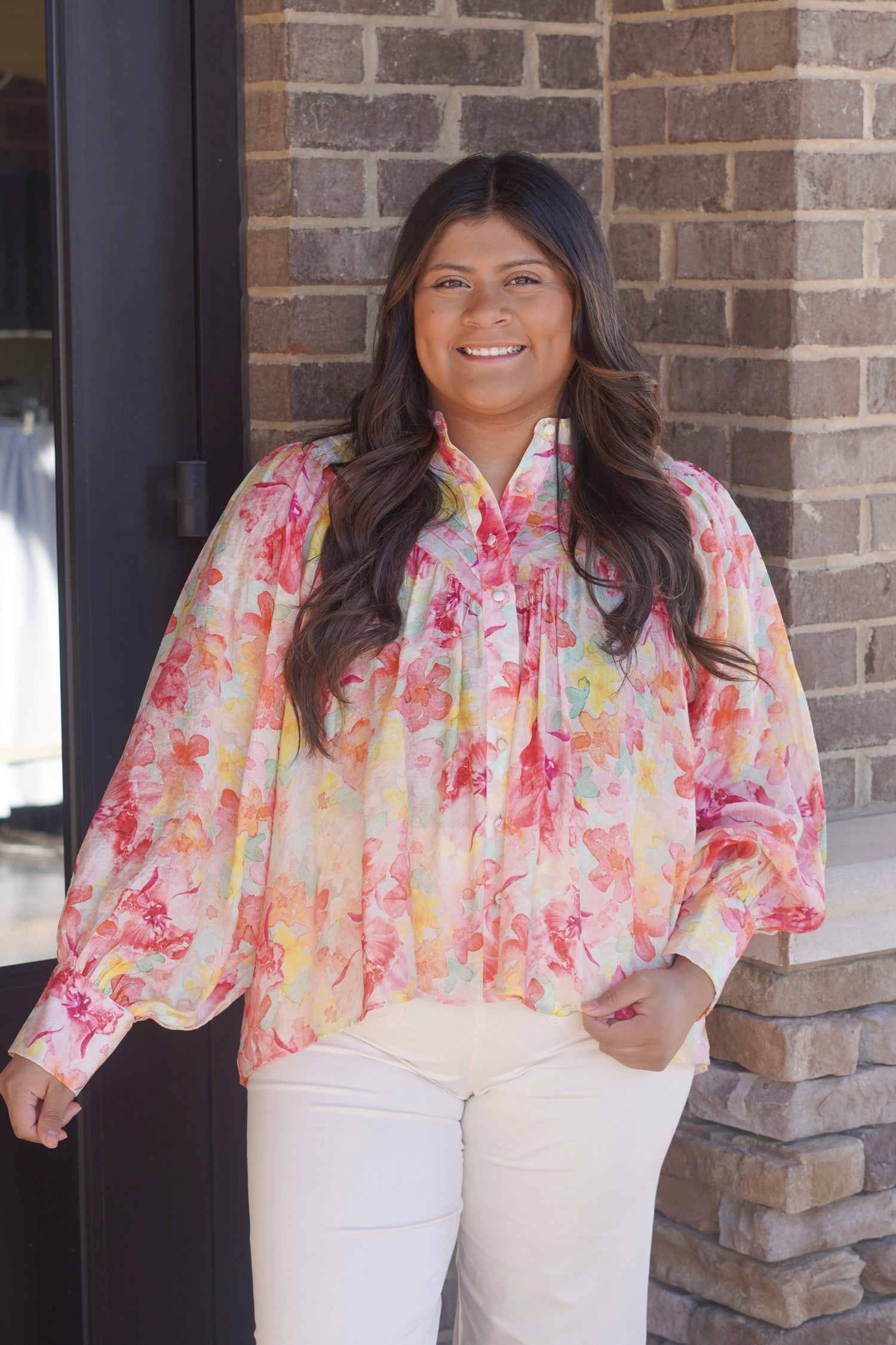 Women wearing a water color floral printed pink, yellow, orange, green blouse, button up, long sleeve cuffed at the wrist, flowy,  paired with some white pants 