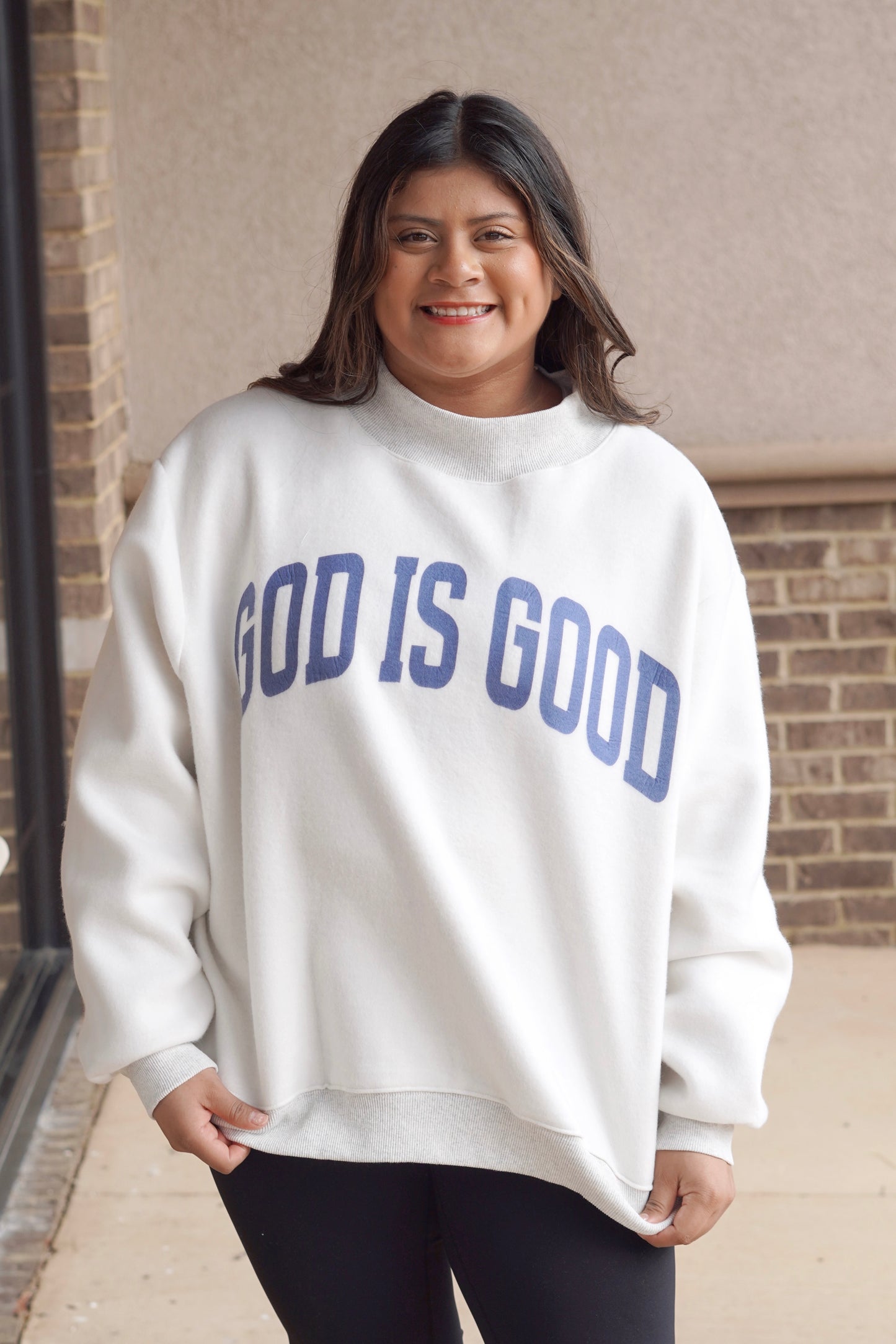 Woman wearing a white oversized mock neck sweatshirt that has "God is Good" written in blue with black leggings. 