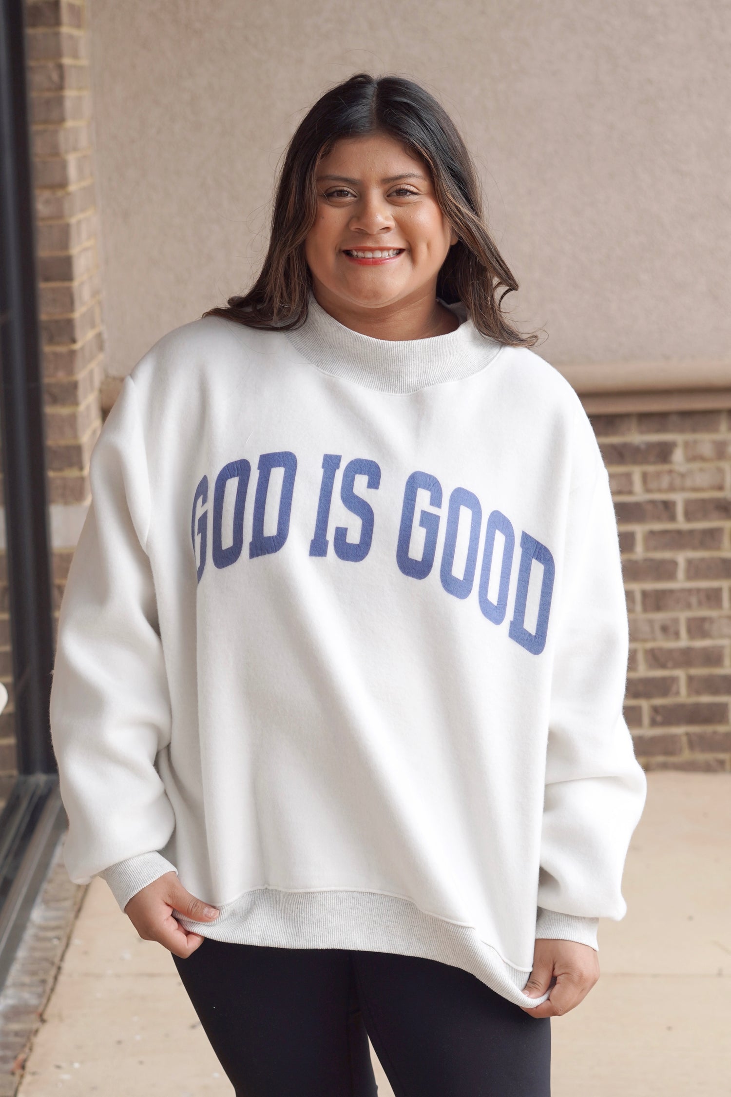 Woman wearing a white oversized mock neck sweatshirt that has "God is Good" written in blue with black leggings. 