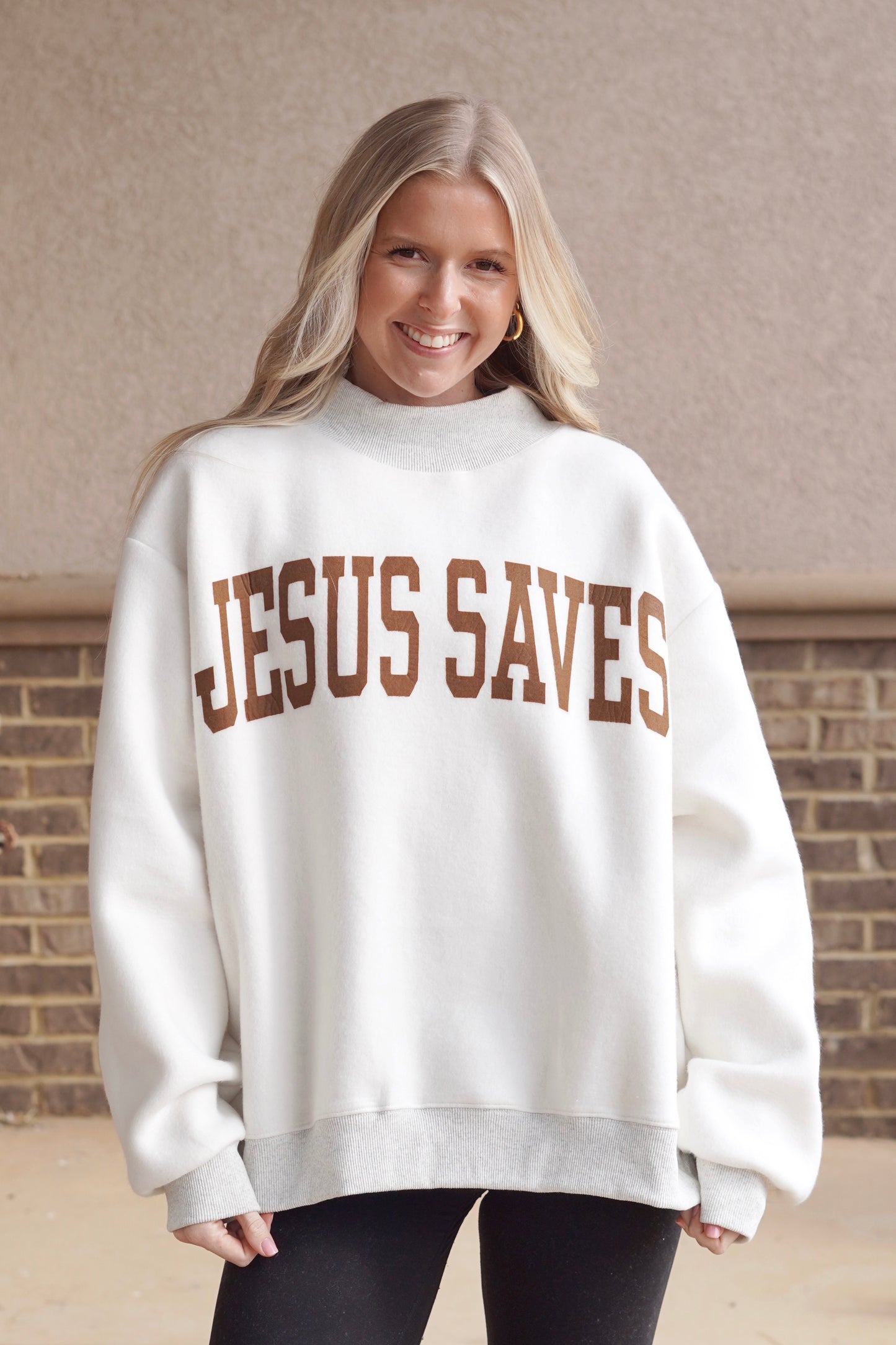 Woman wearing a white oversized mock neck sweatshirt that has "Jesus Saves" written in brown with black leggings. 