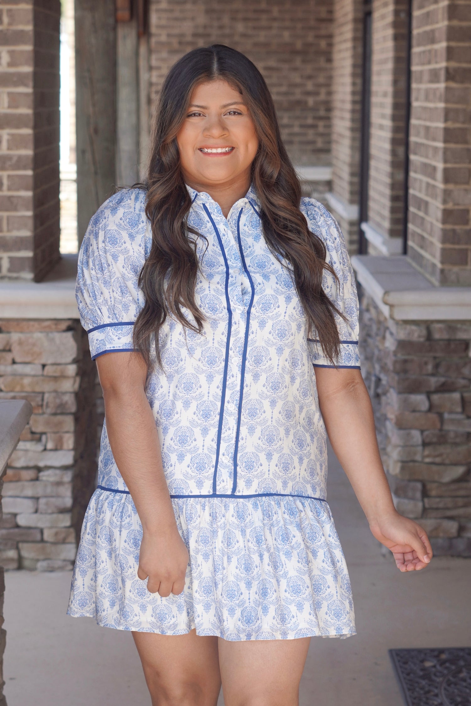  Women wearing a short, collared dress with a blue and white floral pattern, short sleeves and a ruffled hem, with blue trim, buttons going down the middle. 