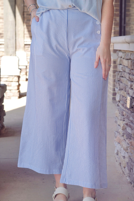 Blue thin striped pants with a white base, wide leg, buttons on the side pocket and elastic waist band 
