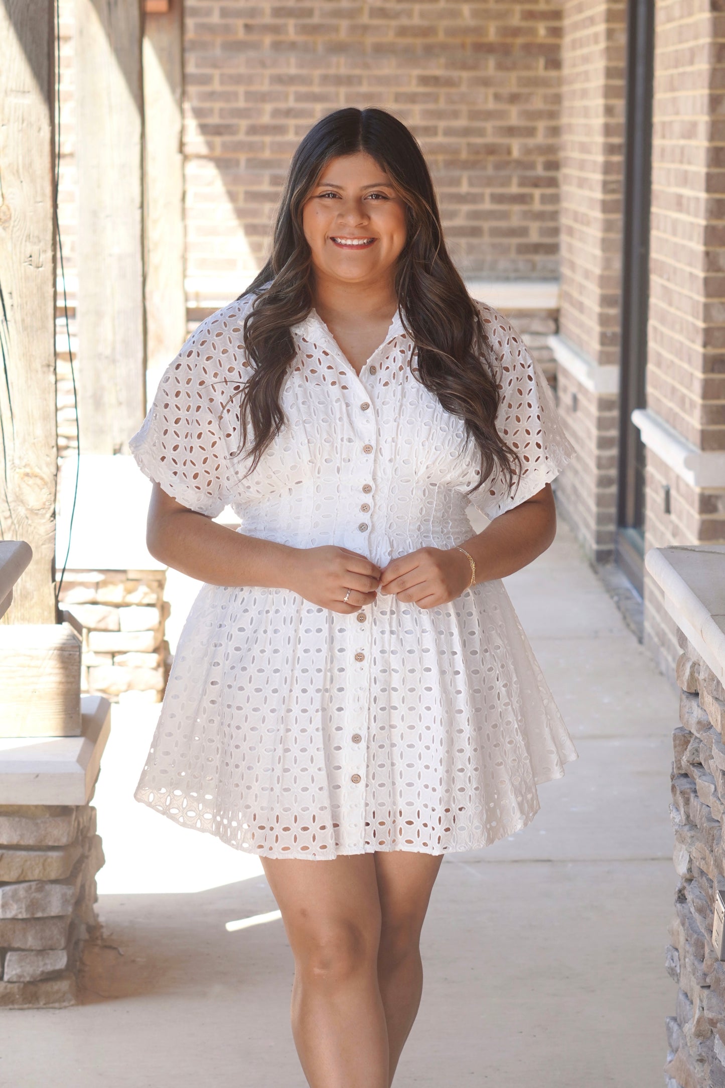 Women wearing a short white eyelet dress with buttons going down the middle, cinch at the waist, short sleeve, collar neckline. 