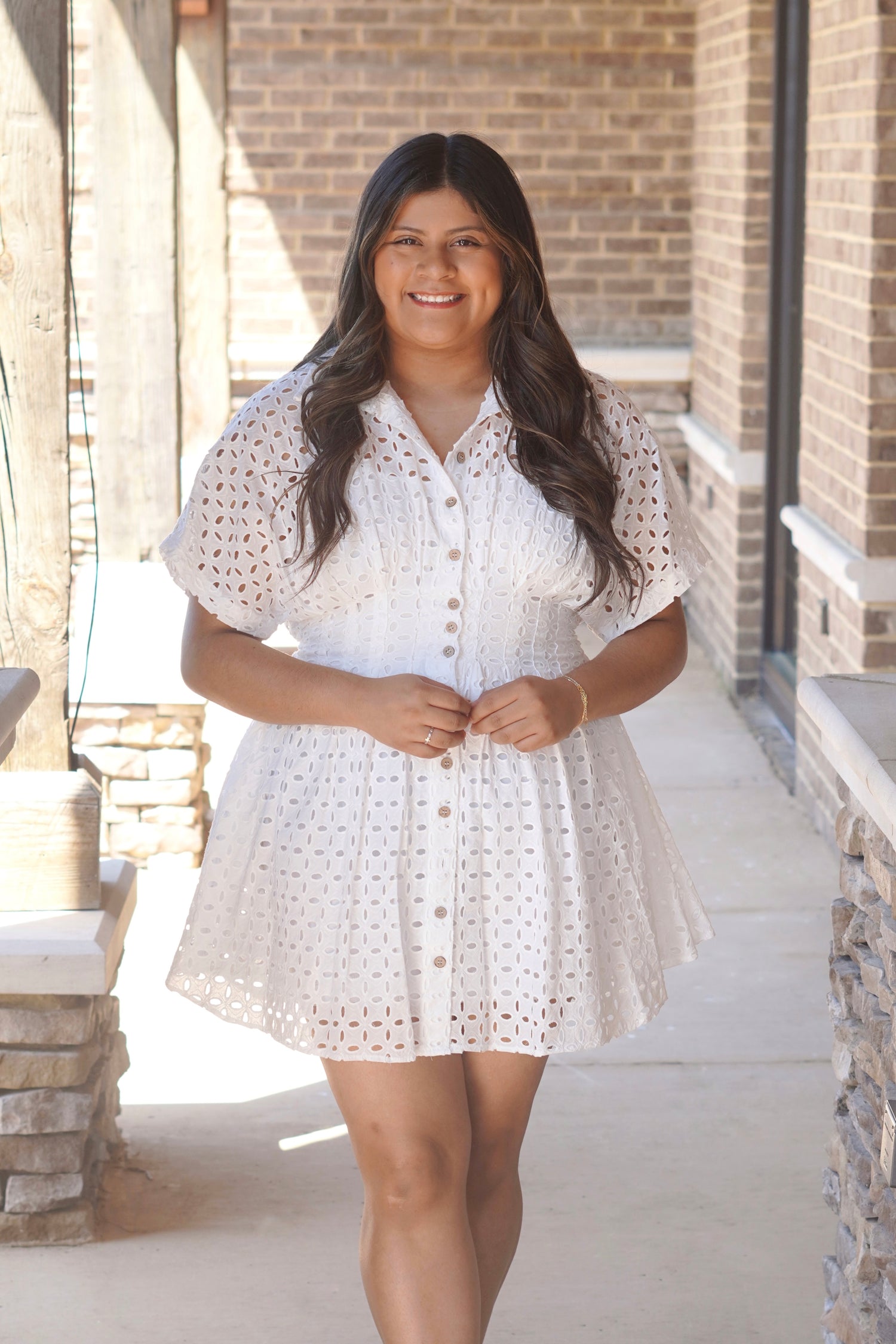 Women wearing a short white eyelet dress with buttons going down the middle, cinch at the waist, short sleeve, collar neckline. 