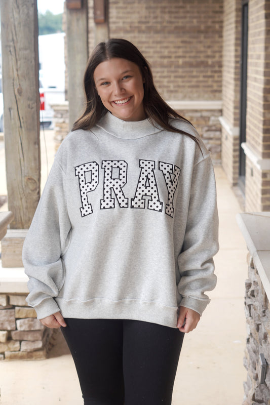 A woman wearing a light gray mock neck sweatshirt with 'PRAY' in black-and-white polka dot block letters across the chest.
