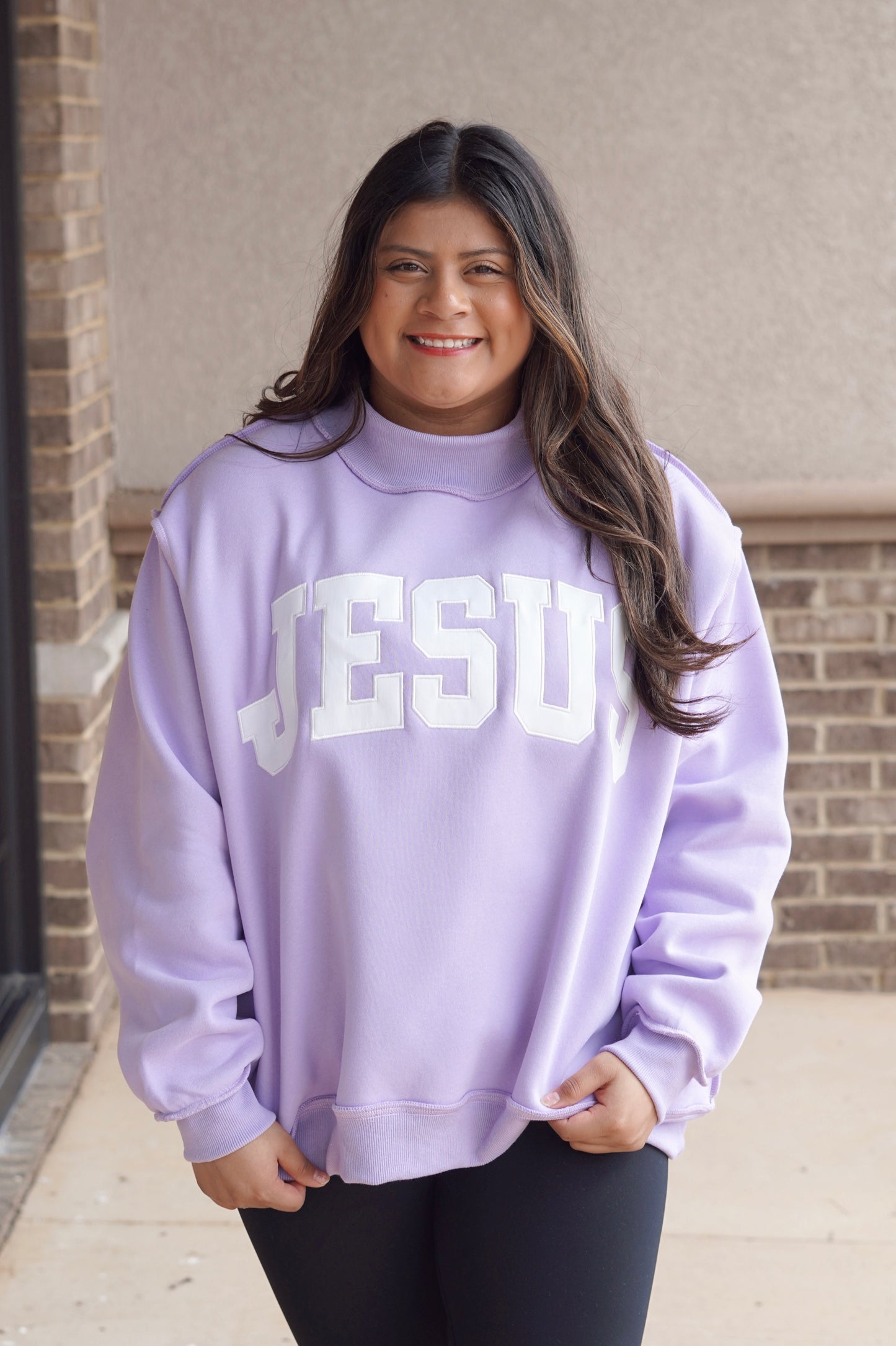 Woman wearing a lavender oversized mock neck sweatshirt that has "Jesus" embroidered in white with black leggings. 