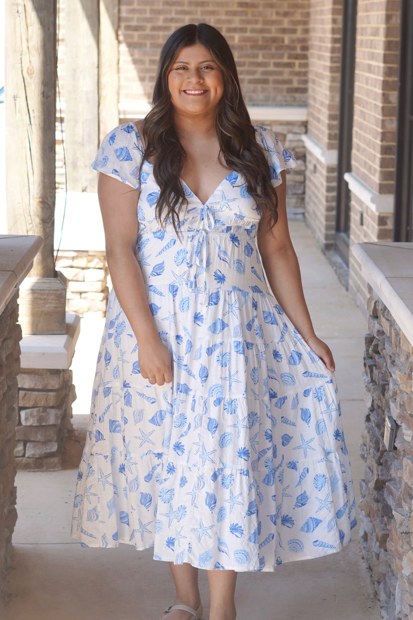 Women wearing a white with blue ocean print dress, with a v neckline, flutter sleeve, midi style. 