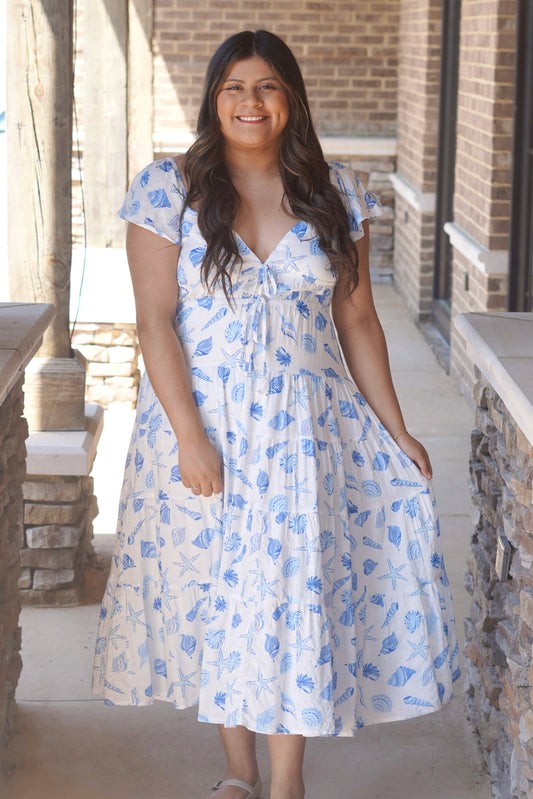 Women wearing a white with blue ocean print dress, with a v neckline, flutter sleeve, midi style.