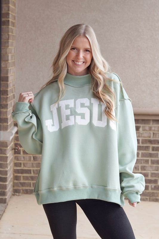 Woman wearing a sage green oversized mock neck sweatshirt that has "Jesus" embroidered in white with black leggings.