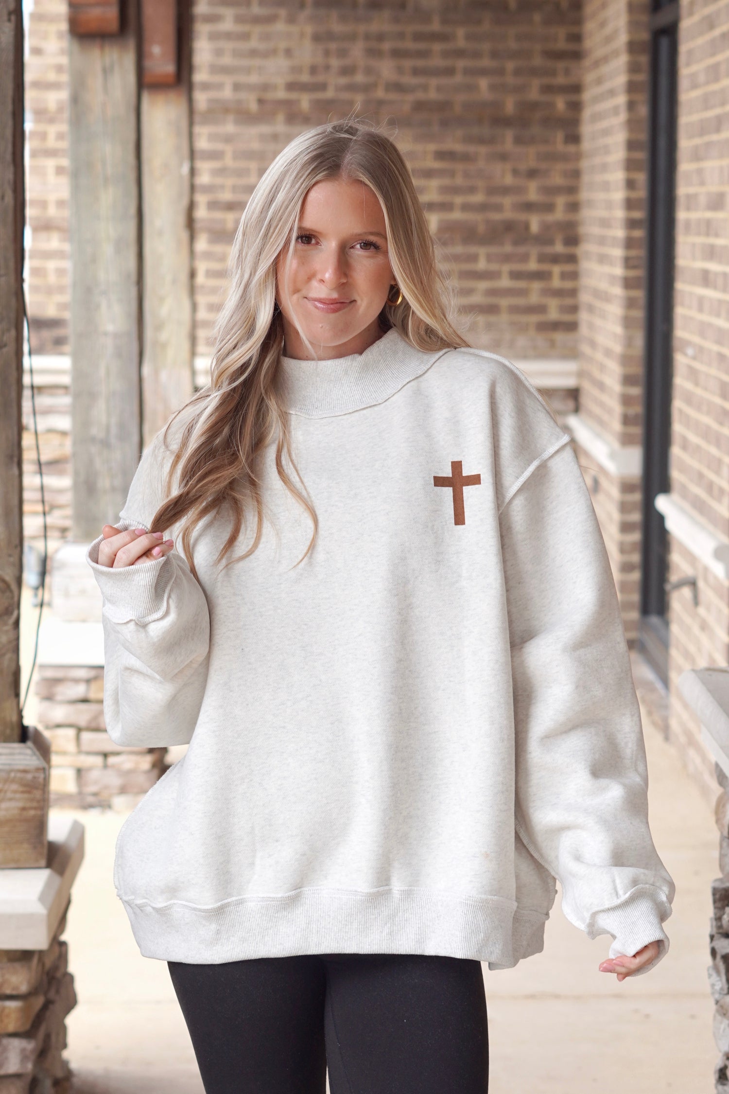 Woman wearing a heather grey oversized mock neck sweatshirt that has a small brown cross on the left side with black leggings. 