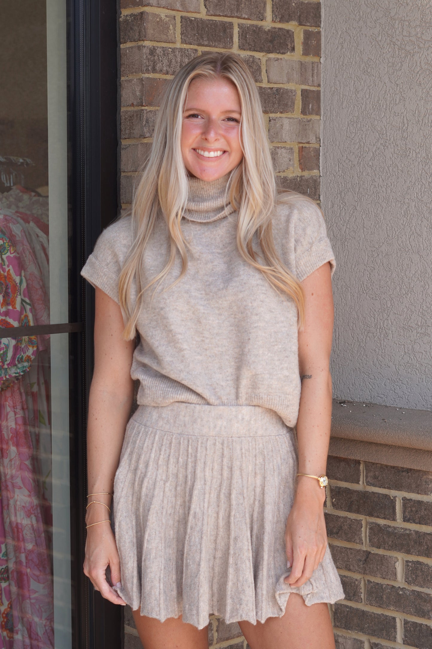 A woman wearing a taupe short-sleeve turtleneck top and matching taupe pleated mini skirt,