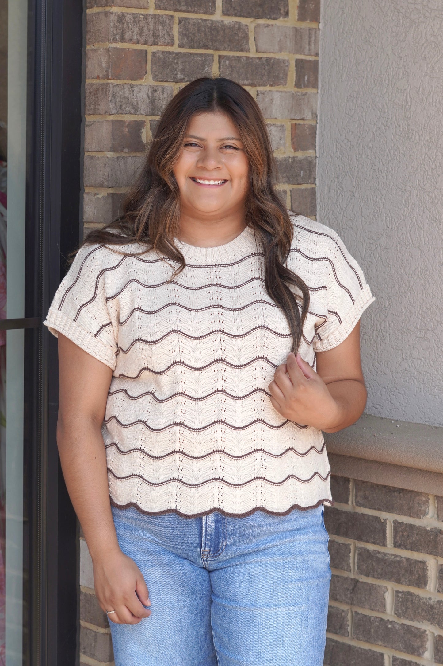 A woman wearing a cream knit short-sleeve top with brown scallop horizontal stripes and light blue jeans, scallop hem. 