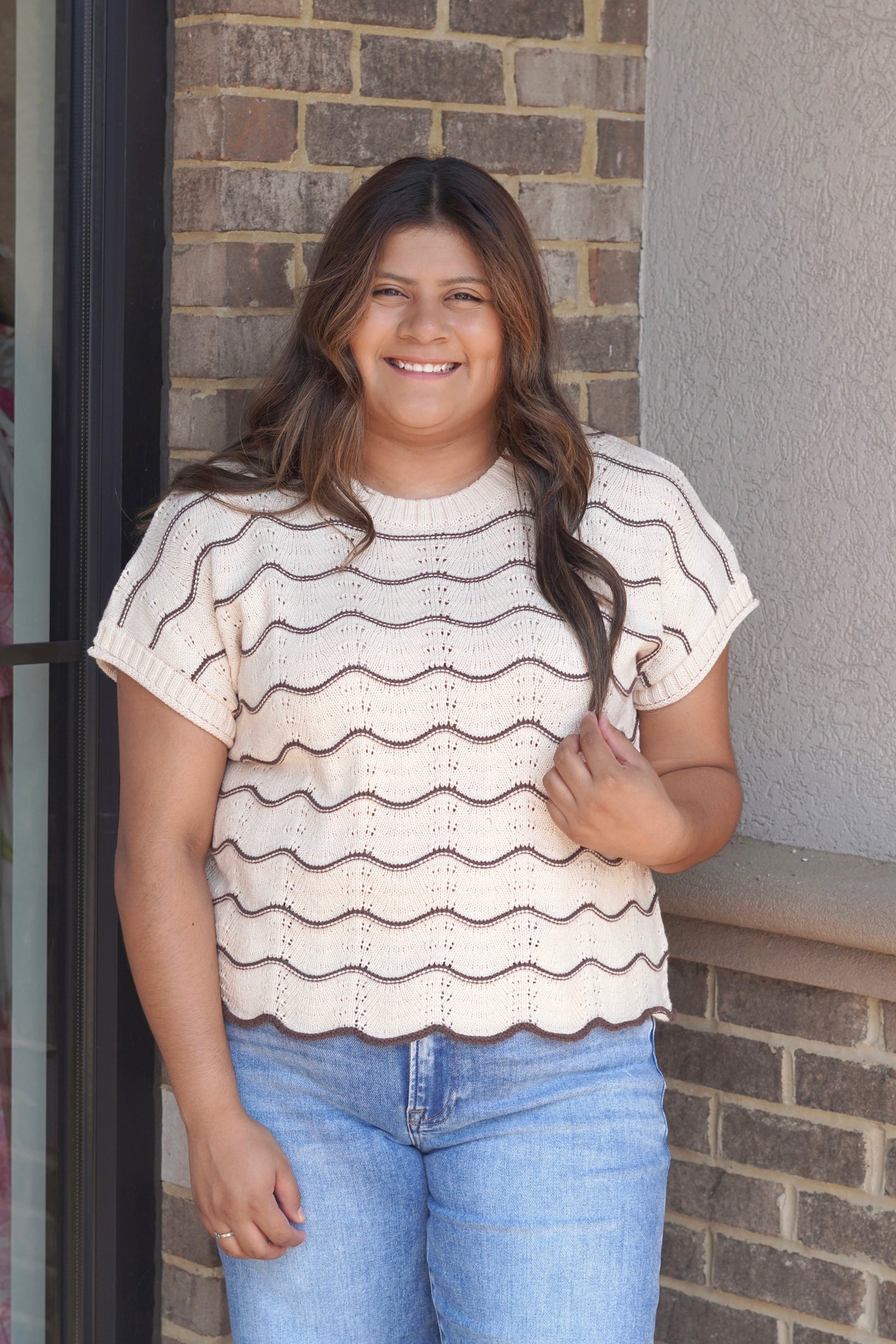 A woman wearing a cream knit short-sleeve top with brown scallop horizontal stripes and light blue jeans, scallop hem. 