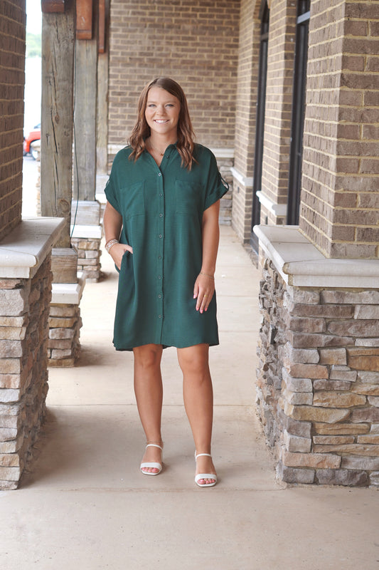 A woman wearing a short-sleeve button up dress in dark green, above the knee length. 