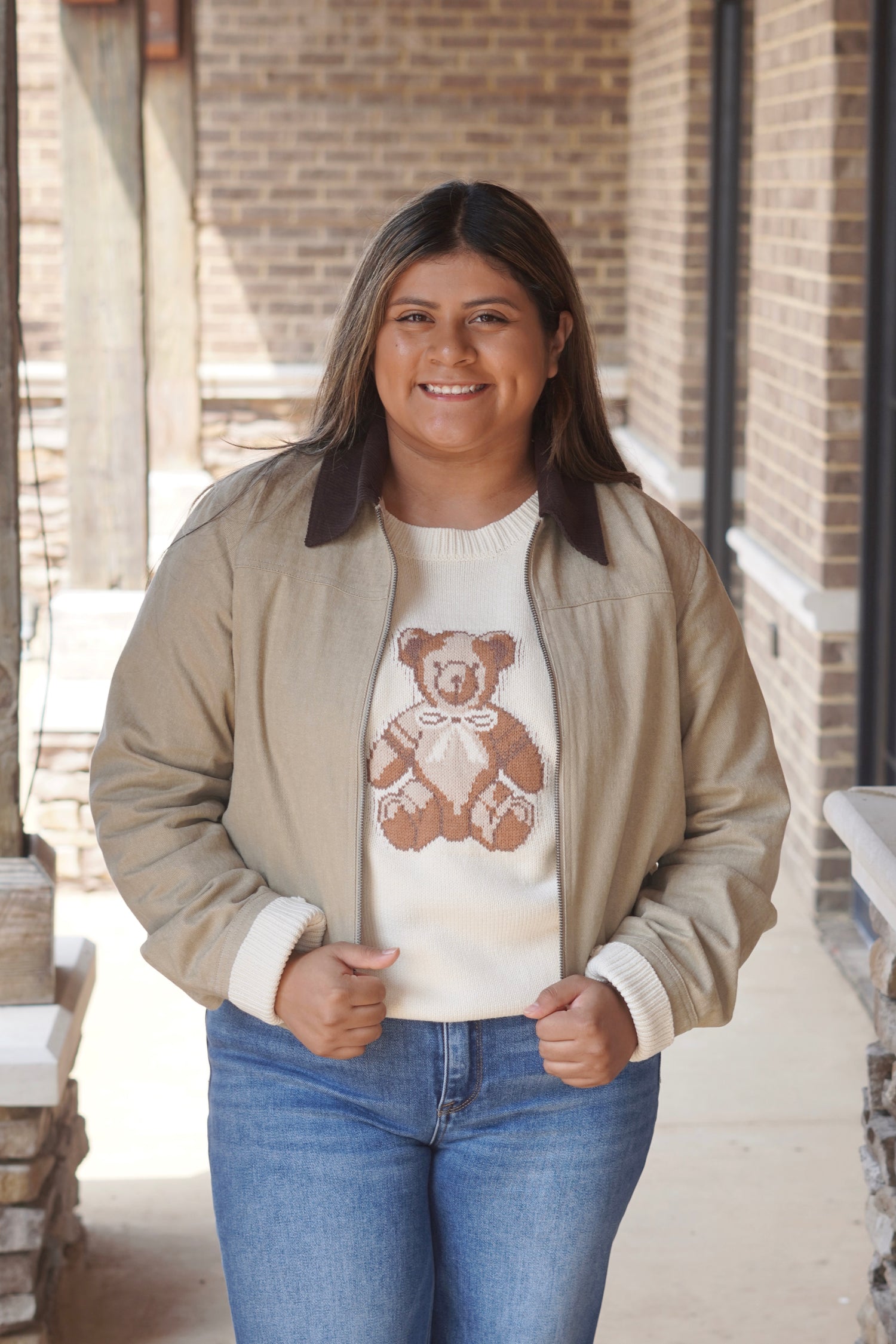 Woman's wearing a cream colored knit sweater with a teddy bear on it paired with a tan colored jacket and medium wash jeans
