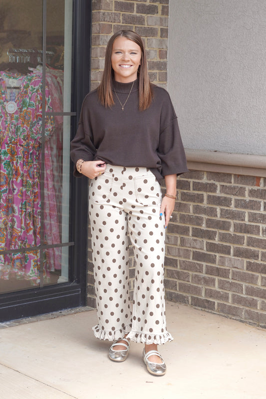 A woman wearing a dark brown long-sleeve top and cream wide-leg pants with brown polka dots and ruffled hems, paired with metallic flats.