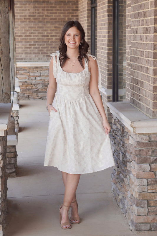woman wearing a cream-colored, knee-length dress with a square neckline and tie straps, floral print 
