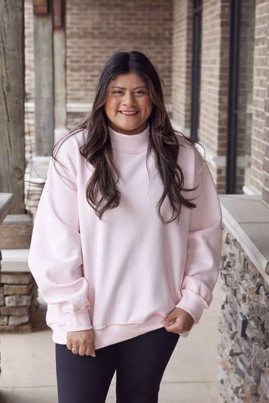 Woman wearing a baby pink oversized mock neck sweatshirt with black leggings.