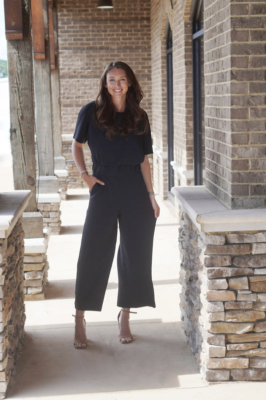 Model wearing a black jumpsuit with a round neckline and cinched waist, as well as a cropped pant leg