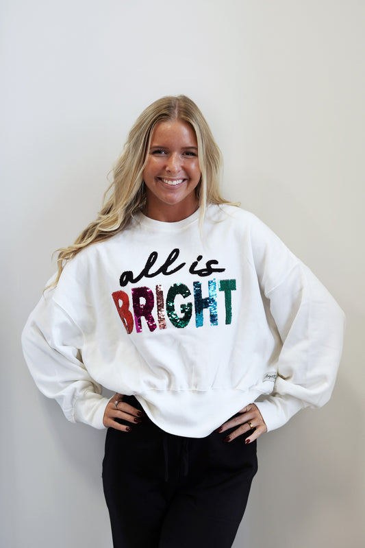 "All is Bright" Script and Sequins Top Crew Neckline Long Cuffed Sleeves Color: Off White "All is" in Black Script Letters "BRIGHT" in Colorful Sequin Letters Ribbed Hem Relaxed Fit Skimmer Length 60% Cotton, 40% Polyester